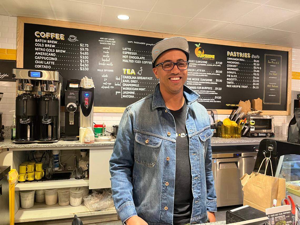 An eye-level, medium shot of a smiling restaurateur wearing a denim jacket, glasses, and a backward baseball cap, standing behind the counter at a coffee shop. Behind, a large chalkboard menu displays various coffee and pastry items.