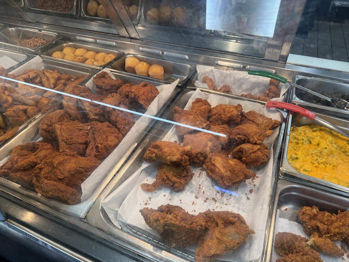 A close-up view looking through a glass sneeze guard at a hot food cafeteria display. Several metal trays contain large, golden-brown pieces of crispy fried chicken on white parchment paper. Background trays hold side items including cornbread muffins, mac and cheese, and potato wedges.