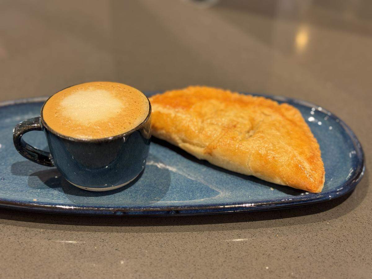 A golden-brown, flaky beef empanada with a crimped edge paired with a small dark blue cup of cortado featuring a smooth layer of espresso foam. Both are served on an oblong blue ceramic platter.
