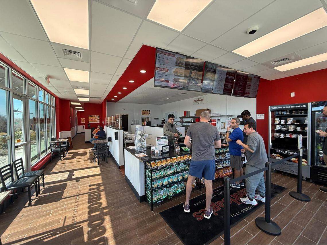 A wide interior shot of the PrimoHoagies restaurant. Customers are standing at the black-topped service counter. The space features red accent walls, wooden-style flooring, digital menu boards hanging from the ceiling, and a refrigerated “Grab and Go” section.