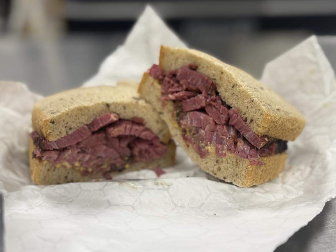 A close-up of a thick pastrami sandwich served on seeded rye bread. The sandwich is sliced in half and resting on white deli paper, revealing layers of thinly sliced, steamed pink meat with a visible spice rub on the edges.