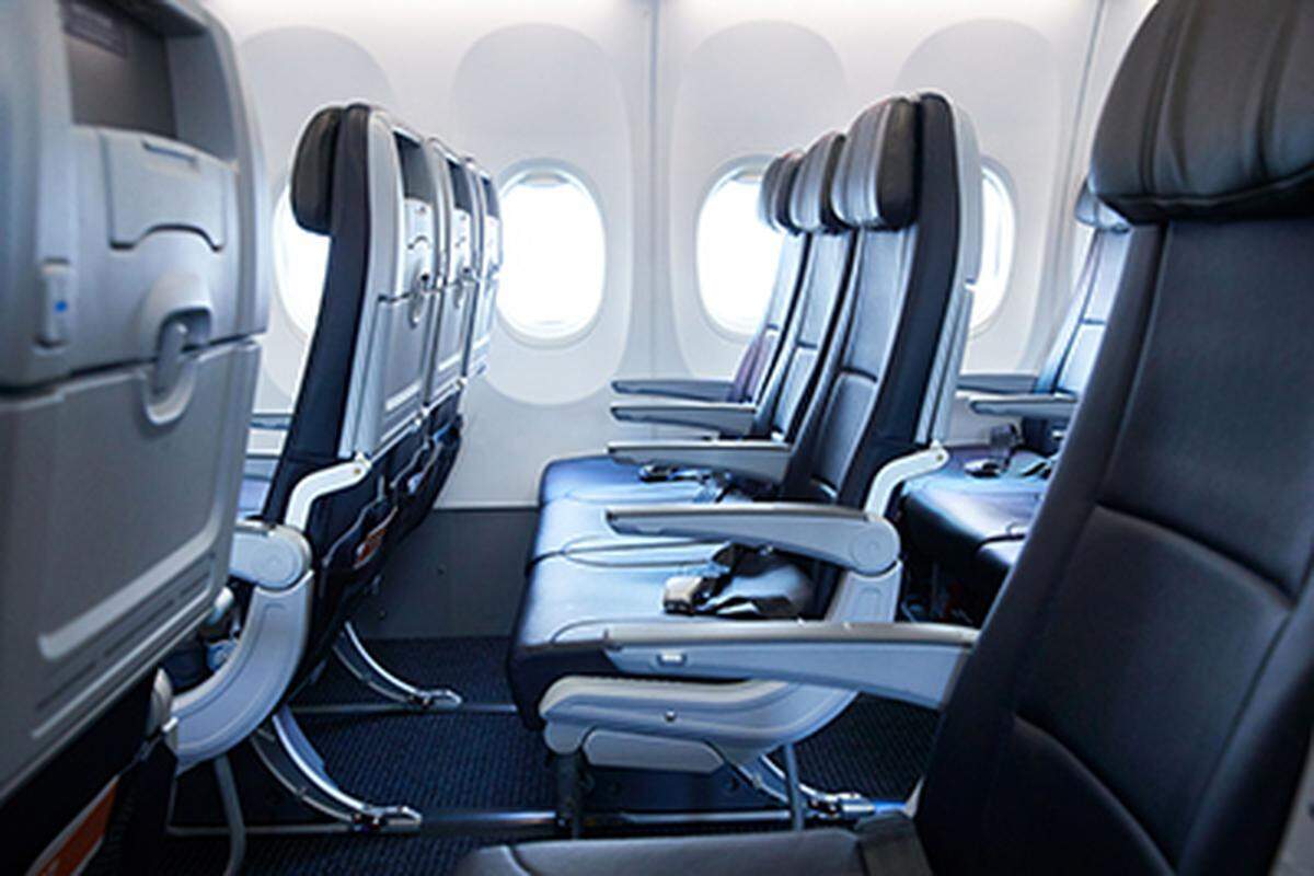 This photo shows the interior of a commercial airplane’s main cabin, featuring a row of dark gray economy seats along the window. The seats are modern with adjustable headrests, and three oval passenger windows are visible in the bright daylight.