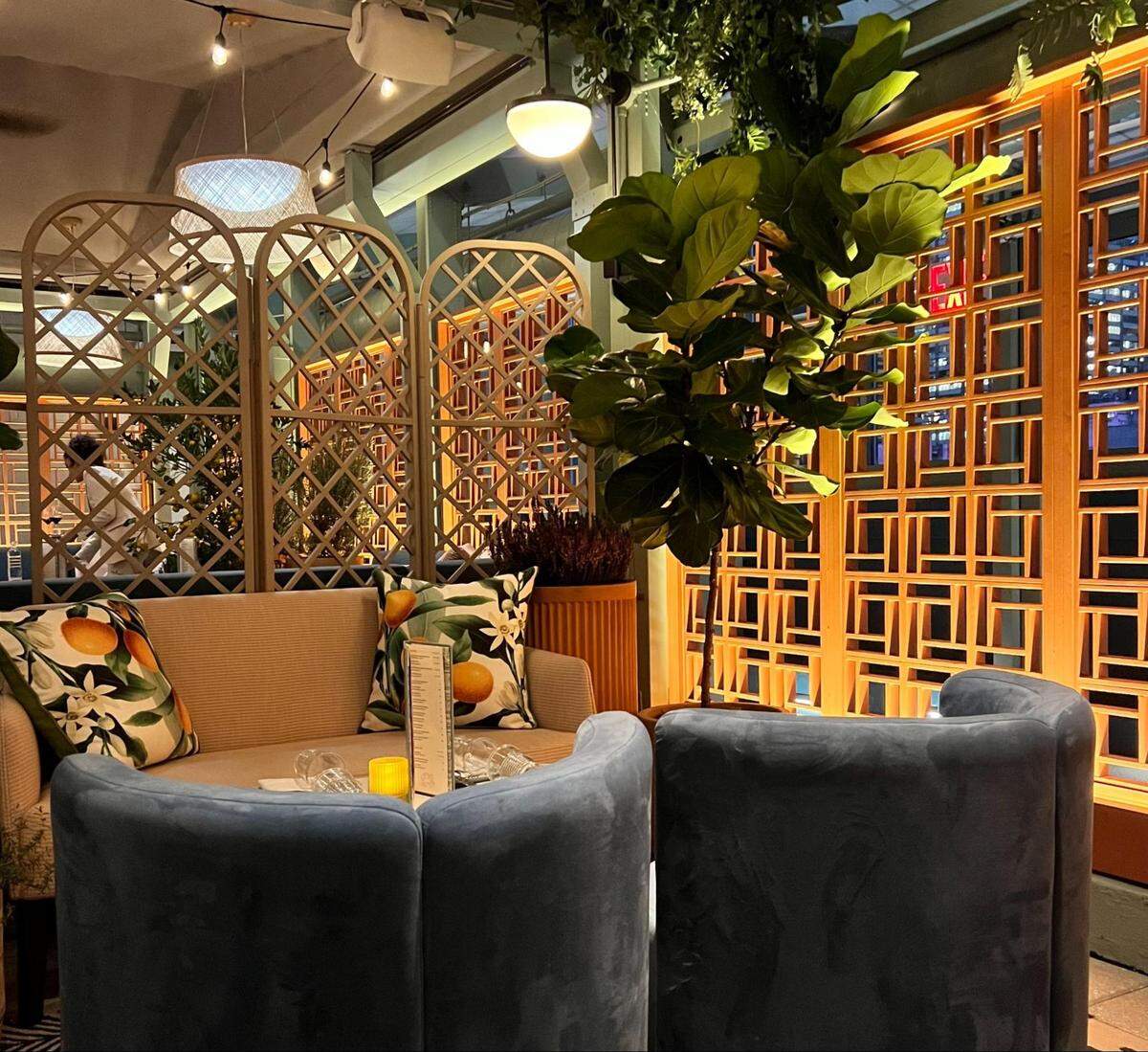 A cozy and stylish lounge area within a restaurant or bar. The space features a small, light-colored sofa with pillows decorated with an orange floral pattern, facing two curved blue armchairs. A small table with a lit candle and glasses sits between the seating. The area is separated by decorative, light-colored wooden screens with geometric lattice patterns. A large fiddle-leaf fig plant stands in a planter, adding a touch of green to the space. The lighting is warm and dim, and the overall atmosphere feels intimate and inviting.