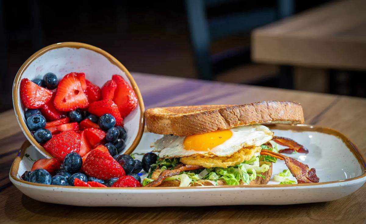A brunch sandwich and a side of fresh fruit served on a white, brown-rimmed plate. The sandwich consists of thick-cut toasted bread layered with shredded lettuce, crispy bacon strips, a fried green tomato, and a fried egg with a bright yellow, runny yolk. On the left side of the plate, a small matching bowl is tilted forward, overflowing with sliced strawberries and fresh blueberries that spill onto the plate.