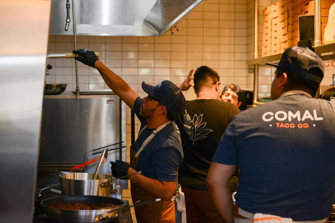 A candid shot inside a busy commercial kitchen with stainless steel surfaces and white subway tile. Several chefs are working; in the foreground, one chef in an apron and cap reaches up to use equipment under the hood, and another chef is seen from the back wearing a shirt with the “COMAL TACO CO.” logo.