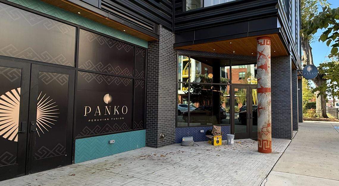 The ground-floor entrance of a new restaurant. Black-framed glass doors and windows display the text “PANKO PERUVIAN FUSION” and a large circular logo. A painted, decorative pillar is on the right, near some buckets and stacked tiles on the patio.