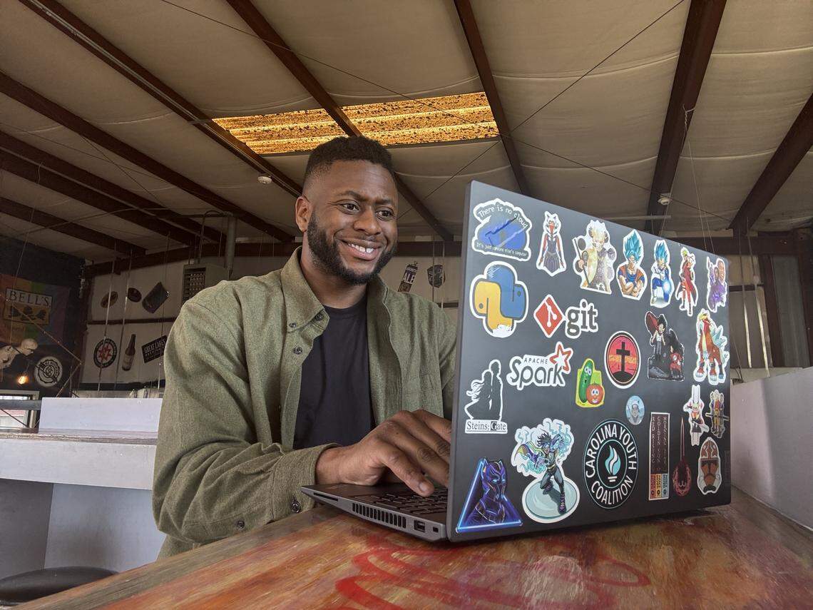 A smiling person with a beard, wearing a green button-down shirt over a black t-shirt, sits at a wooden table in an industrial-style indoor space, typing on a dark laptop heavily decorated with various stickers.