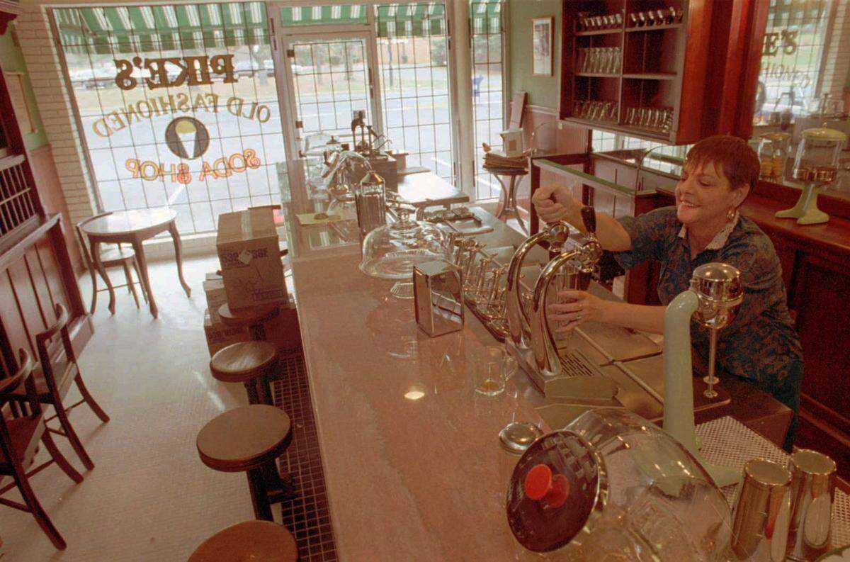 A 1994 high-angle photograph of a woman with short hair smiling as she pulls a chrome soda fountain tap behind a long, polished counter. The interior belongs to “Pike’s Old Fashioned Soda Shop,” visible through the reverse lettering on the large front window. The shop features wooden stools, vintage glass cake domes, and warm wooden cabinetry, all illuminated by bright natural light coming through the storefront.