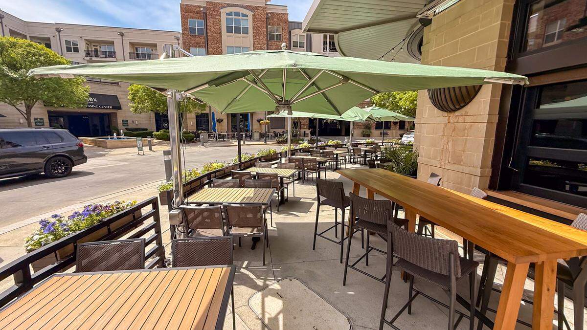 A sunny outdoor dining area located on a paved sidewalk. The patio is filled with wooden-slatted tables and grey mesh chairs, all shaded by large, sage green rectangular umbrellas. A long, high-top wooden communal table sits near the building’s stone facade. Black metal planters filled with purple and yellow flowers line the perimeter, separating the patio from the street and nearby apartment buildings.