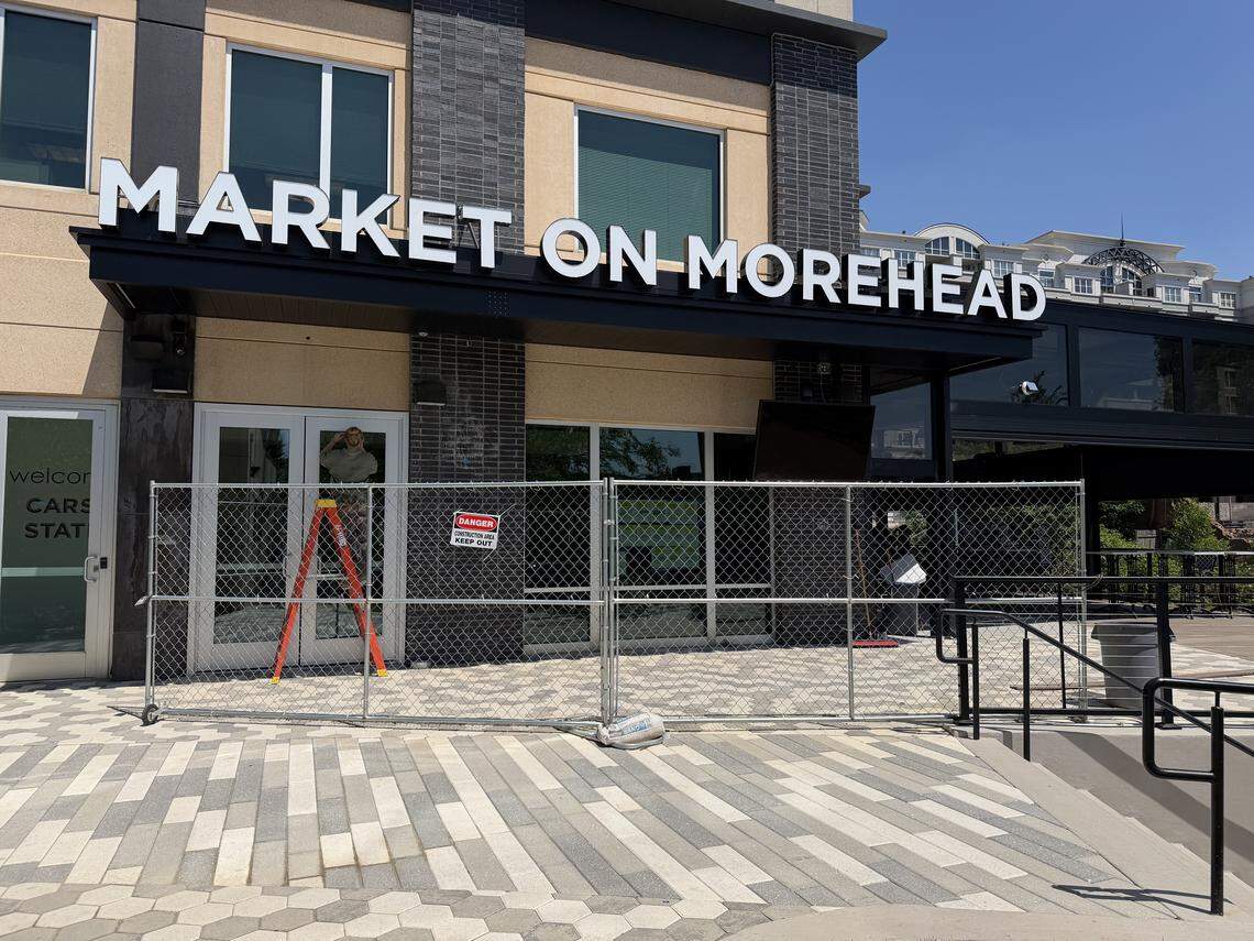 A street-level view of the “Market on Morehead” storefront featuring a large black awning and white signage. A construction fence and ladder are visible in front of the glass entrance.