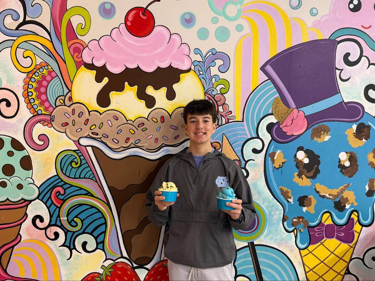A teenager in a grey UNC windbreaker smiles while holding a small blue cup of pale yellow ice cream with a black plastic spoon. The teenager is standing in front of a colorful wall mural featuring a giant ice cream sundae with a cherry on top.