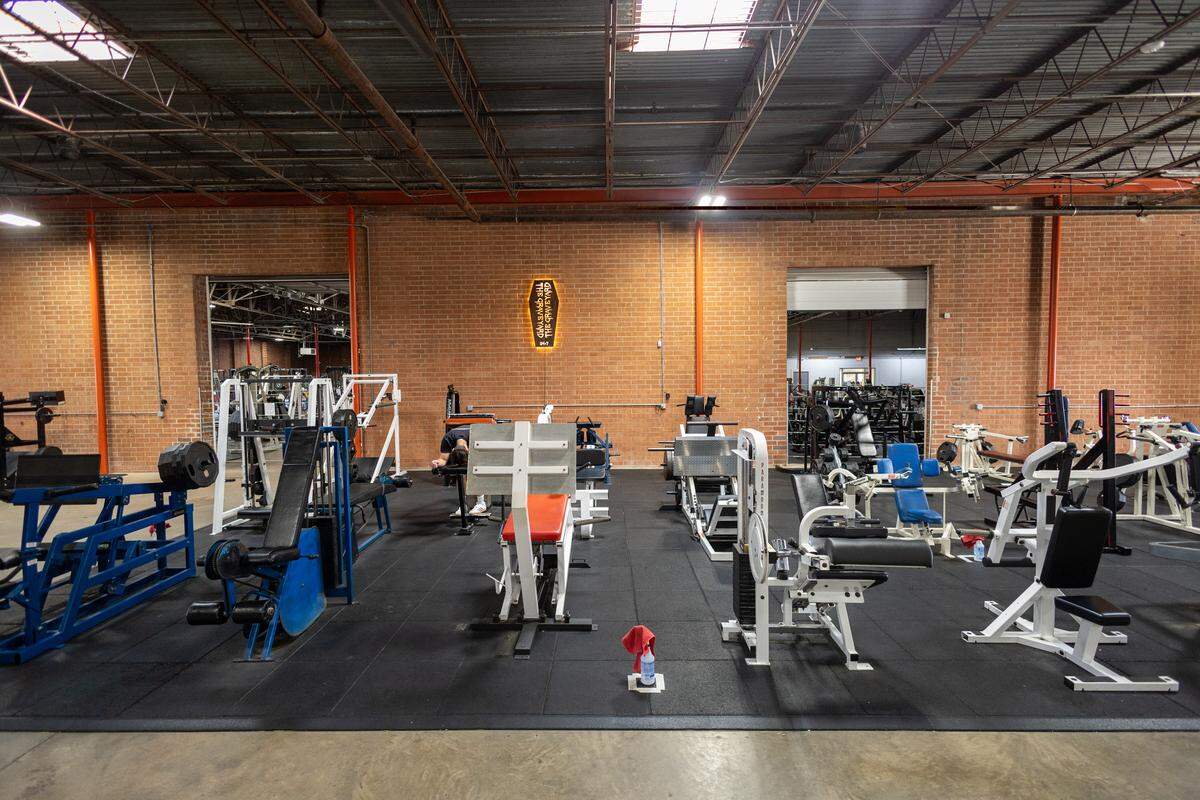 Symmetrical view of a gym interior with a brick wall background, showcasing multiple white and blue vintage weight machines. A lit, coffin-shaped sign hangs in the center above the equipment.