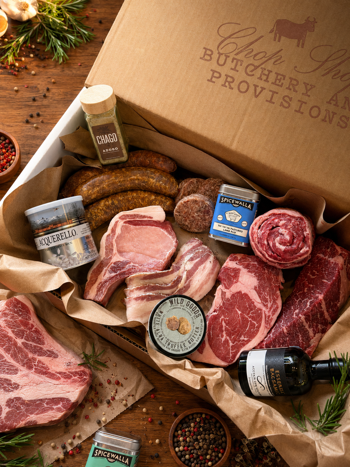 An overhead shot of a curated gourmet meat and provisions box from “Chop Shop Butchery and Provisions,” packed in brown butcher paper inside a cardboard box. The box features an assortment of raw, high-quality meats including thick bone-in pork chops, heavily marbled ribeye steaks, a rolled roast, sausage links, and bacon strips. Interspersed with the meats are premium pantry items: a tin of Spicewalla spice blend, a jar of Chago Adobo seasoning, a container of Acquerello rice, a small jar of Wild Goods White Alba Truffle Salt, and a bottle of Essenza Balsamic vinegar. The scene is set on a warm wooden surface accented with loose peppercorns, garlic bulbs, and fresh rosemary sprigs.