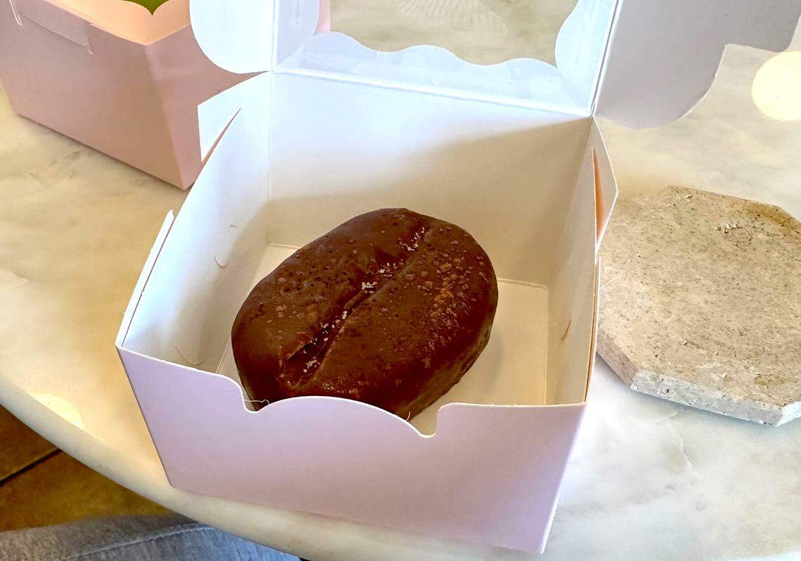 A brown pastry shaped like a giant coffee bean sits inside an open pink pastry box on a white marble table.