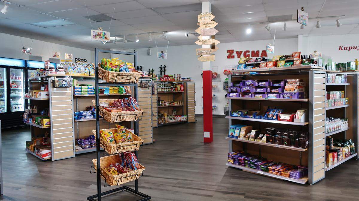 A view down an aisle at Zygma market, with shelves heavily stocked with European chocolates and candies, including many varieties of Milka chocolate bars. A stand with baskets of snacks is in the foreground.