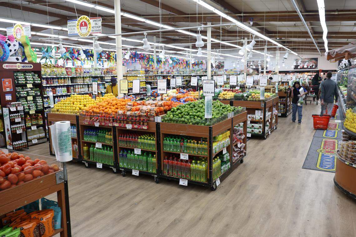 Interior view of a grocery store aisle centered on large wooden display tables piled high with fresh lemons, oranges, and limes. Bottles of juices are stacked underneath the produce. Shoppers are visible in the background.