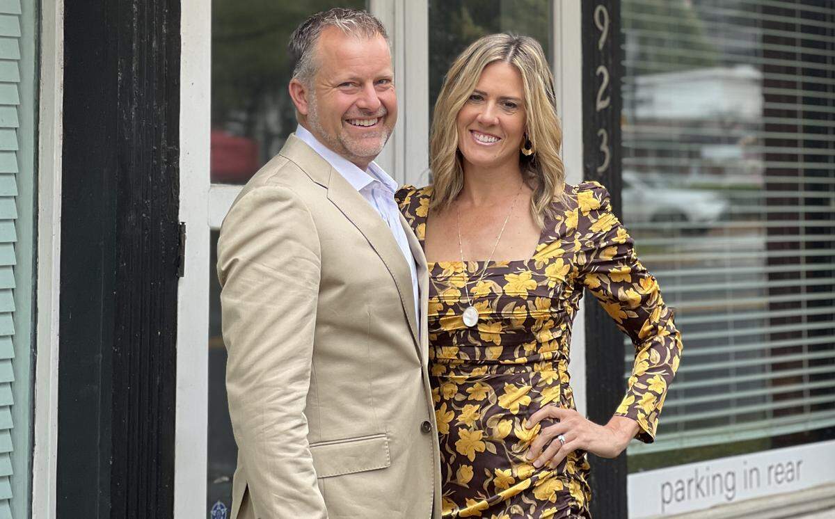 A portrait of a smiling couple, standing closely together in front of a glass storefront. The person on the left is wearing a tan blazer and light blue shirt; the one on the right is wearing a dark brown dress with a yellow floral pattern. The building’s address “1923” is partially visible on the black door frame behind them.