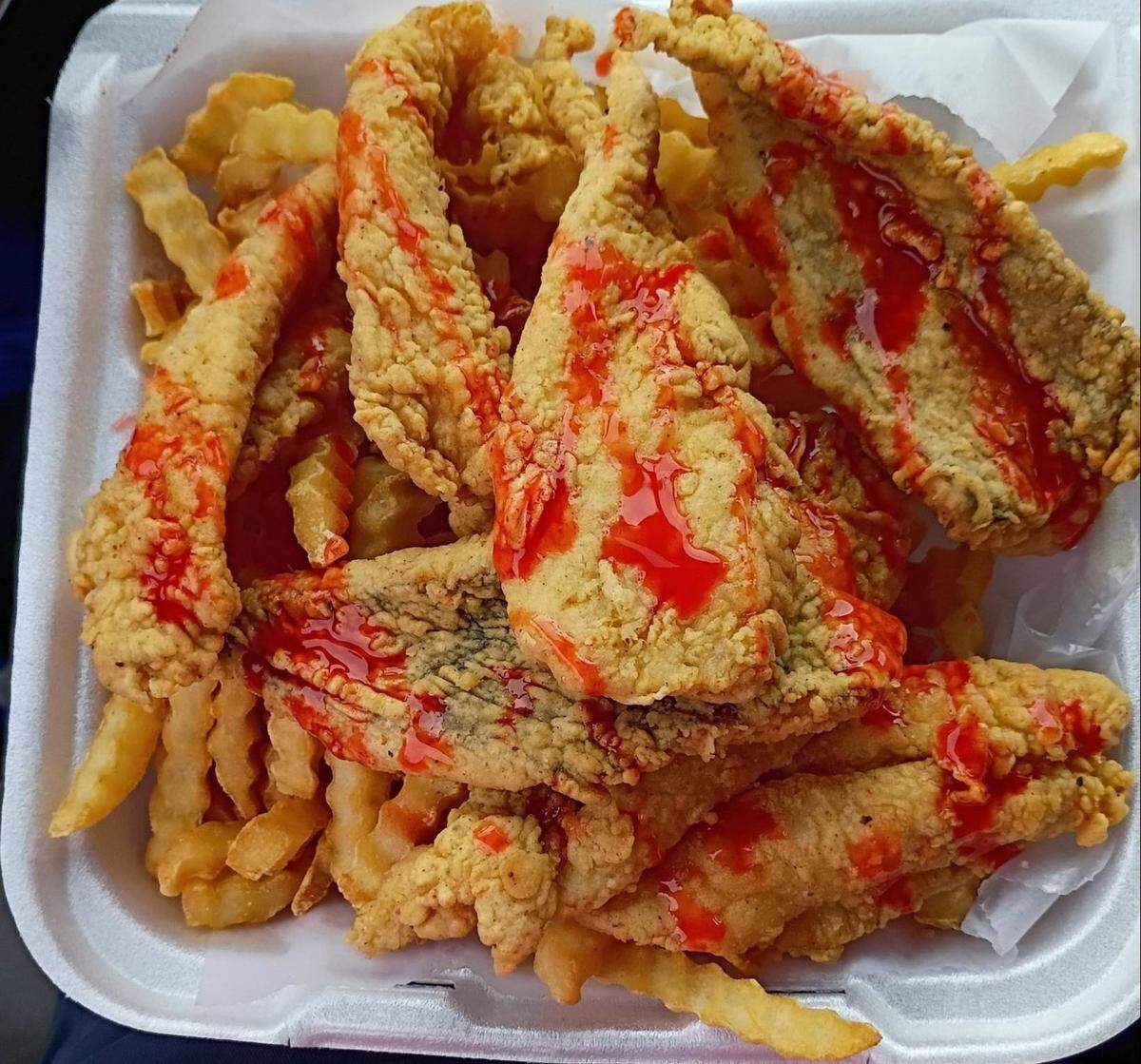A top-down, close-up shot shows a white Styrofoam takeout container overflowing with a classic “fried fish and chips” meal. The container is packed with a large serving of crinkle-cut French fries forming a base for several large, golden-brown fried fish fillets. The fish has a thick, textured cornmeal breading, suggesting a crunchy exterior. A bright red mild or hot sauce is drizzled generously across the top of the fish and fries in a zigzag pattern. The entire meal sits on a small sheet of white wax paper inside the container.