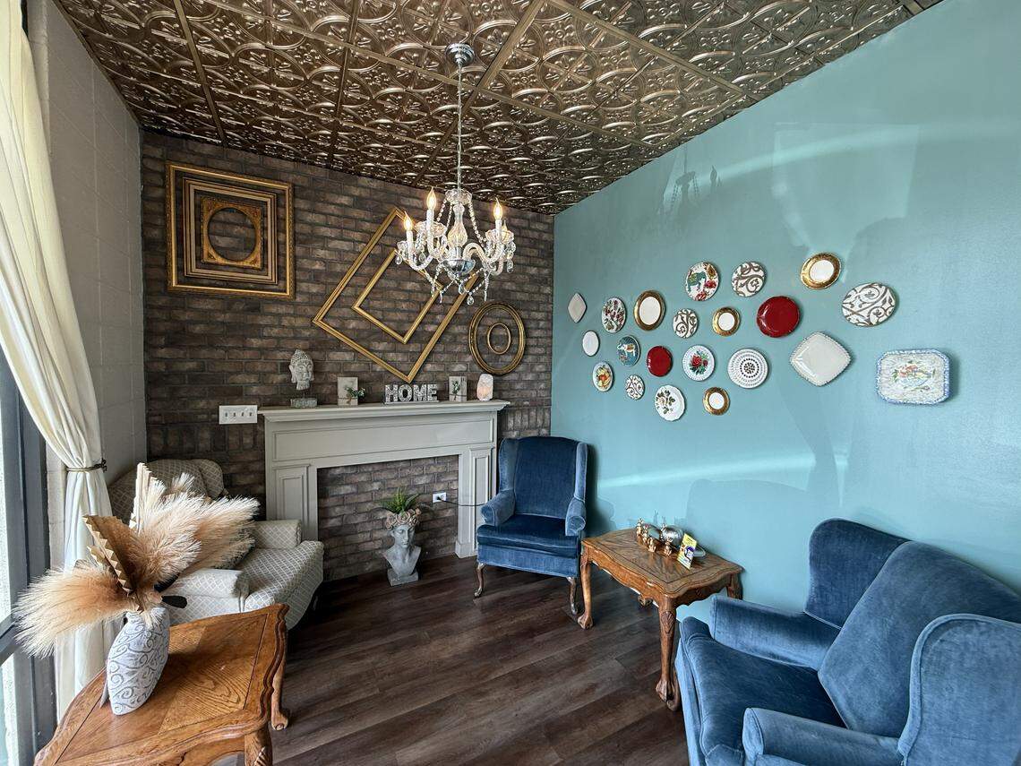 A cozy interior seating area featuring a mix of vintage and eclectic decor. There is a faux brick wall with a white mantelpiece, a teal accent wall adorned with various decorative plates, two blue velvet armchairs, and a metallic patterned ceiling with a crystal chandelier.