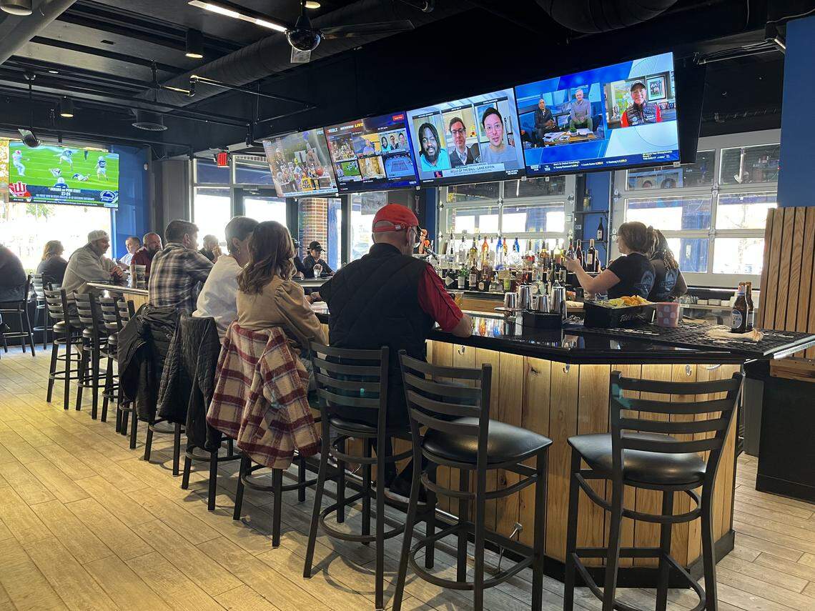 An eye-level, wide shot of a sports bar interior, showing several patrons seated on high-top stools along a long, dark wooden bar. A bartender is visible on the right, and a long row of multiple television screens is mounted overhead, all displaying different programs.