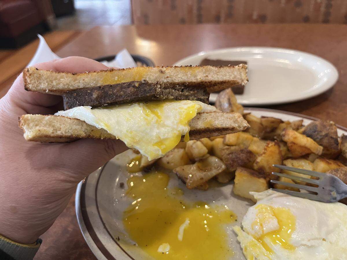 A hand holds up a breakfast sandwich made with toasted white bread, a dark rectangular slice of fried liver mush, and a fried egg with a runny yolk, with excess yolk dripping onto the plate below.