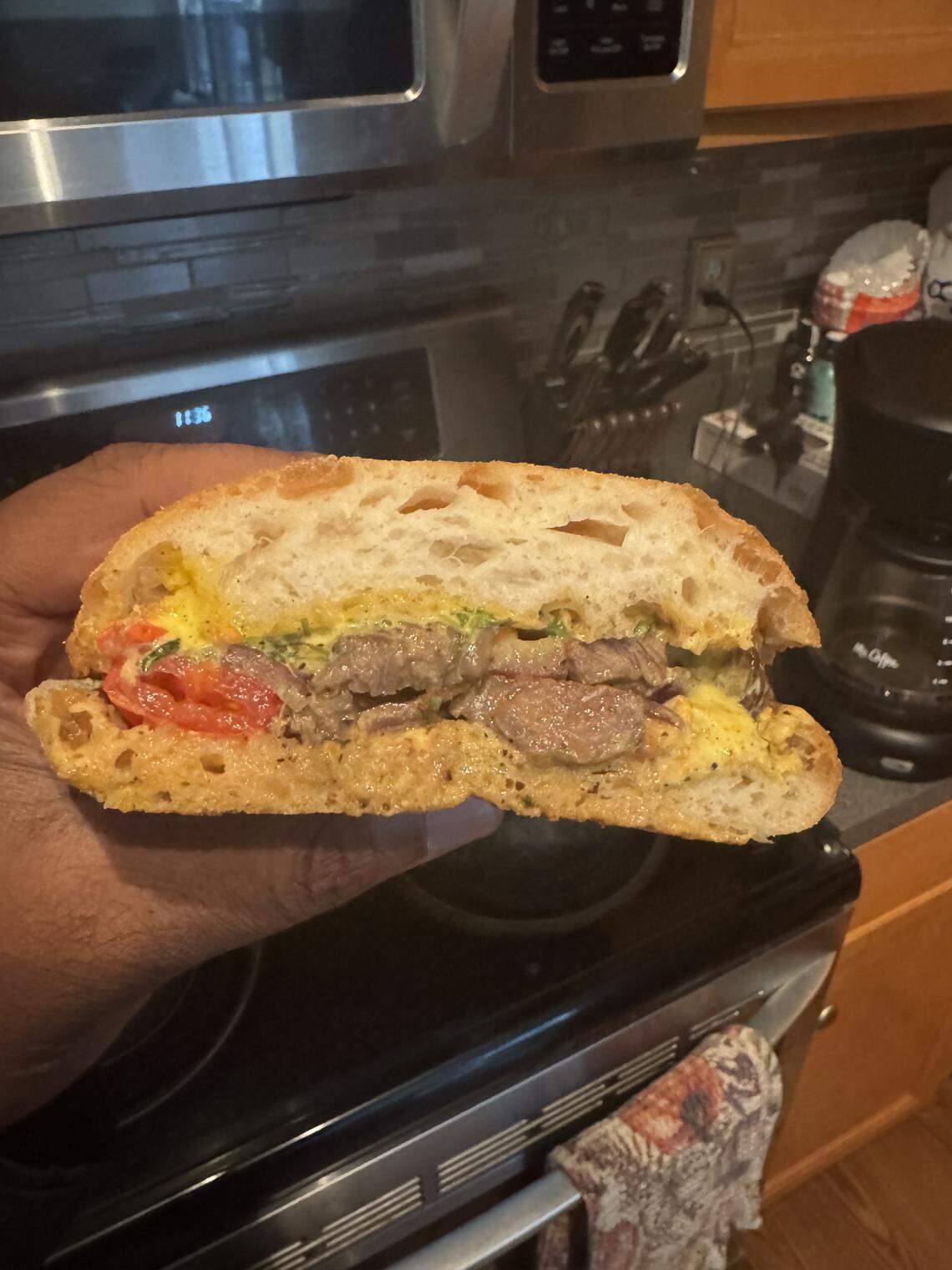 A person’s hand holds a sliced steak sandwich, revealing the filling. The sandwich is made with a crusty bread and appears to contain sliced beef, a red sauce or tomatoes, and a yellow condiment like mustard. The photo is taken in a kitchen with a stove and a backsplash in the background.