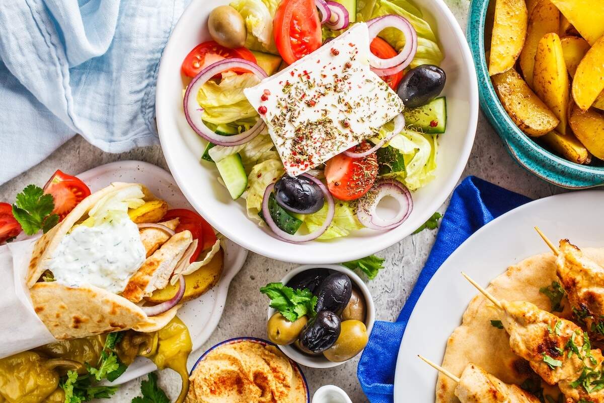 A high-angle, overhead shot of a vibrant Greek food spread on a light gray surface. At the top center sits a white bowl filled with Greek salad containing lettuce, tomatoes, cucumbers, red onions, olives, and a large, thick rectangle of feta cheese seasoned with dried herbs. Surrounding the salad is a chicken gyro wrap drizzled with tzatziki sauce, a plate of grilled chicken souvlaki skewers on flatbread, a bowl of golden roasted potato wedges, a small dish of hummus dusted with paprika, and a small bowl of mixed green and black olives.