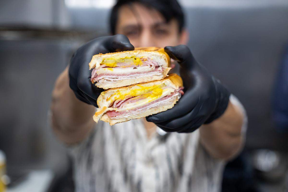 A close-up, shallow-focus shot of a person’s hands in black gloves, holding up a pressed Cuban sandwich. The sandwich is cut in half, prominently displaying its layers of ham, melted white cheese, pickles, and yellow mustard. The person holding it is blurred in the background.