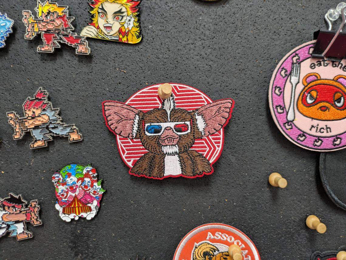 A close-up shot of an embroidered patch featuring Gizmo, the Mogwai from Gremlins, wearing red and blue 3D glasses. The patch has a red and white circular background and is pinned by a small wooden peg to a dark, textured surface. It is surrounded by other pop culture patches and enamel pins, including a Demon Slayer (Rengoku) pin, several pixel-art character pins, and a pink circular patch to the right featuring Tom Nook from Animal Crossing with the text “Eat the rich.”