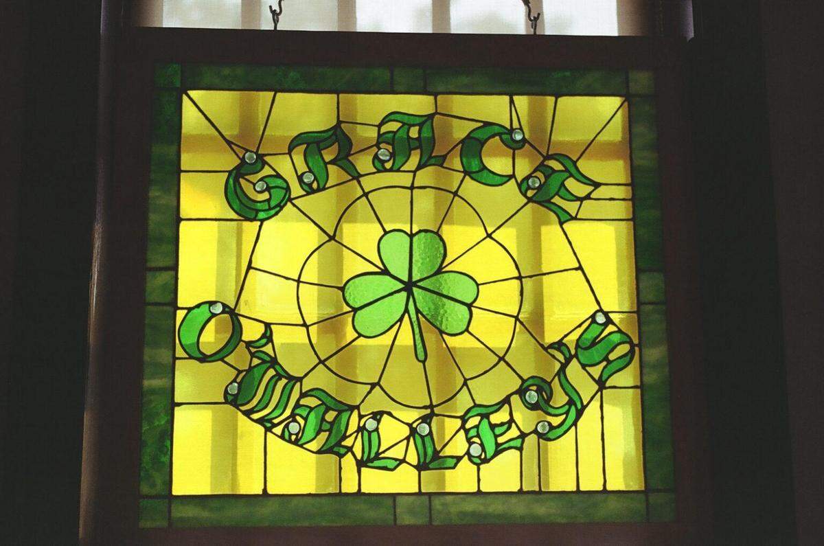 A close-up shot of a square, backlit stained glass window. The design features a large central shamrock surrounded by the name “GRACE O’MALLEY’S” in green Celtic lettering, set within a vibrant yellow and green background.