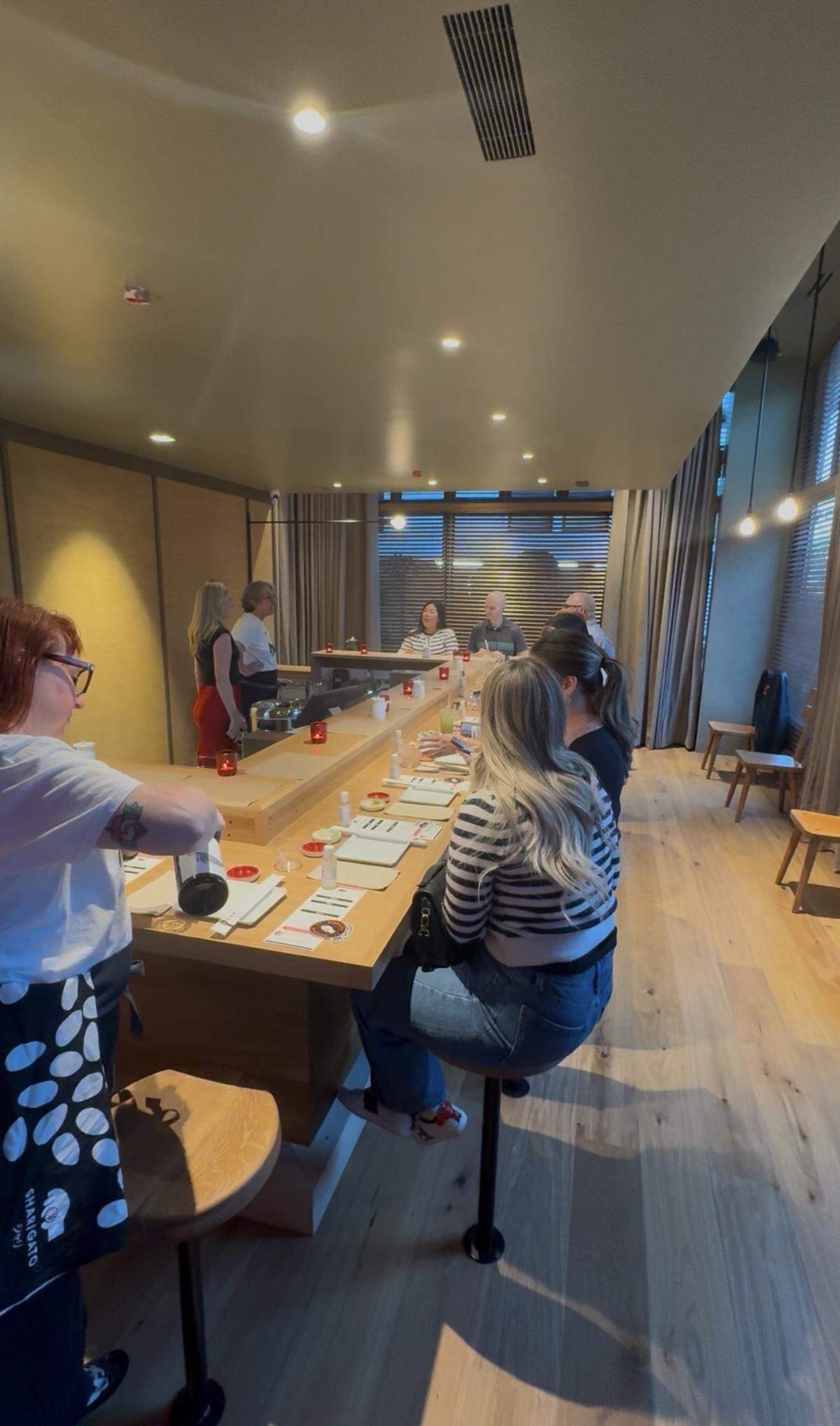 A high-angle, wide shot of the interior of a modern sushi hand roll bar. Patrons sit on tall stools along a sleek, L-shaped light wood counter. The room features minimalist decor with warm overhead lighting, large windows with horizontal blinds, and smooth wooden floors. Staff members are seen behind the counter preparing drinks and serving guests.