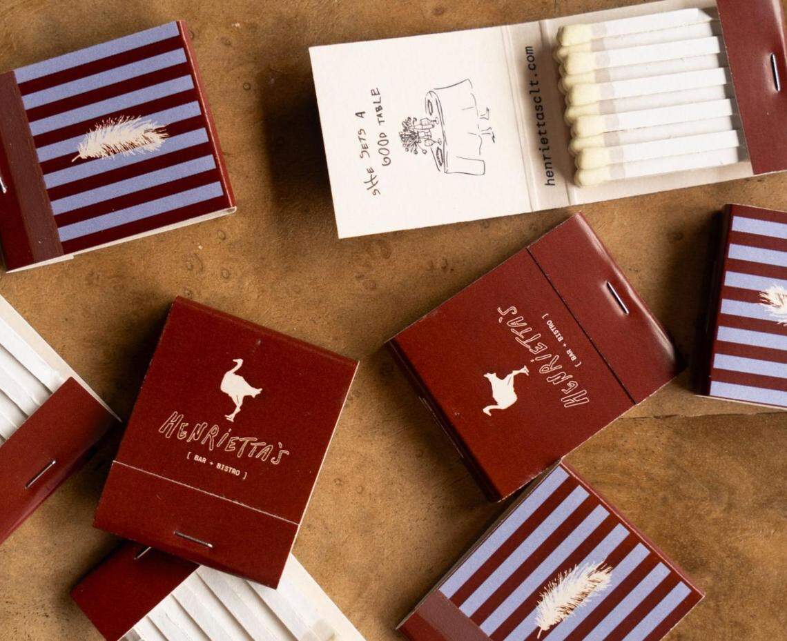An overhead, artfully arranged photo of several custom matchbooks from “Henrietta’s [ Bar • Bistro ]” scattered on a warm brown surface. Some matchbooks are solid maroon with a white ostrich logo, while others have a blue and maroon striped design with a white feather. One matchbook is open, revealing the matches and an illustrated inside flap that reads “SHE SETS A GOOD TABLE” with the website henriettasclt.com.