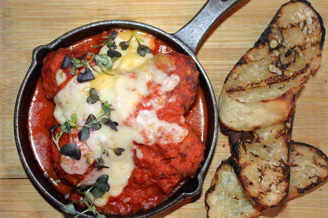 A close-up, top-down shot of a dish of meatballs in a cast-iron skillet. The large meatballs are covered in a rich red tomato sauce and a generous layer of melted white cheese. They are garnished with fresh microgreens. To the right of the skillet, several slices of toasted, crusty bread with visible grill marks are stacked on a wooden cutting board.