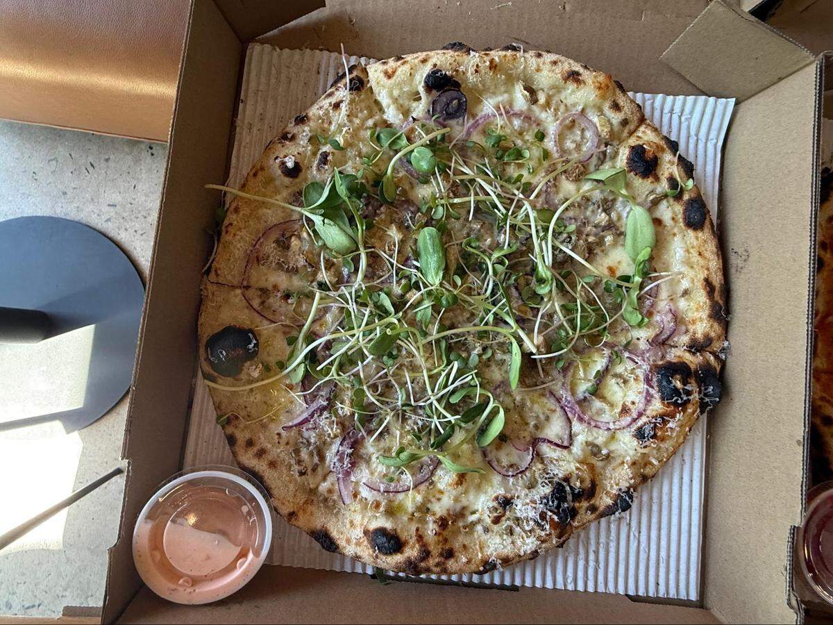 An overhead, close-up shot of an artisan-style pizza in an open brown cardboard box. The pizza has a white sauce base, a bubbly, charred sourdough crust, and is topped with melted cheese, red onion slices, and a generous pile of fresh green microgreens in the center. A small plastic container of a pinkish-orange dipping sauce is in the bottom left corner.