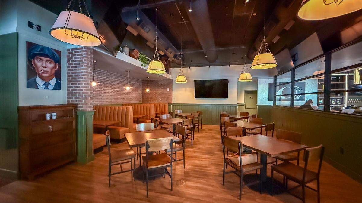 A wide-angle view of a modern Irish pub interior. The room features warm wood flooring, dark ceilings with industrial piping, and a mix of seating including tan leather tufted booths along a brick wall and freestanding wooden tables with dark metal chairs. Large, cream-colored drum pendant lights hang from the ceiling, casting a warm glow. A stylized portrait of Cillian Murphy as Thomas Shelby from Peaky Blinders hangs on a sage green wall to the left.