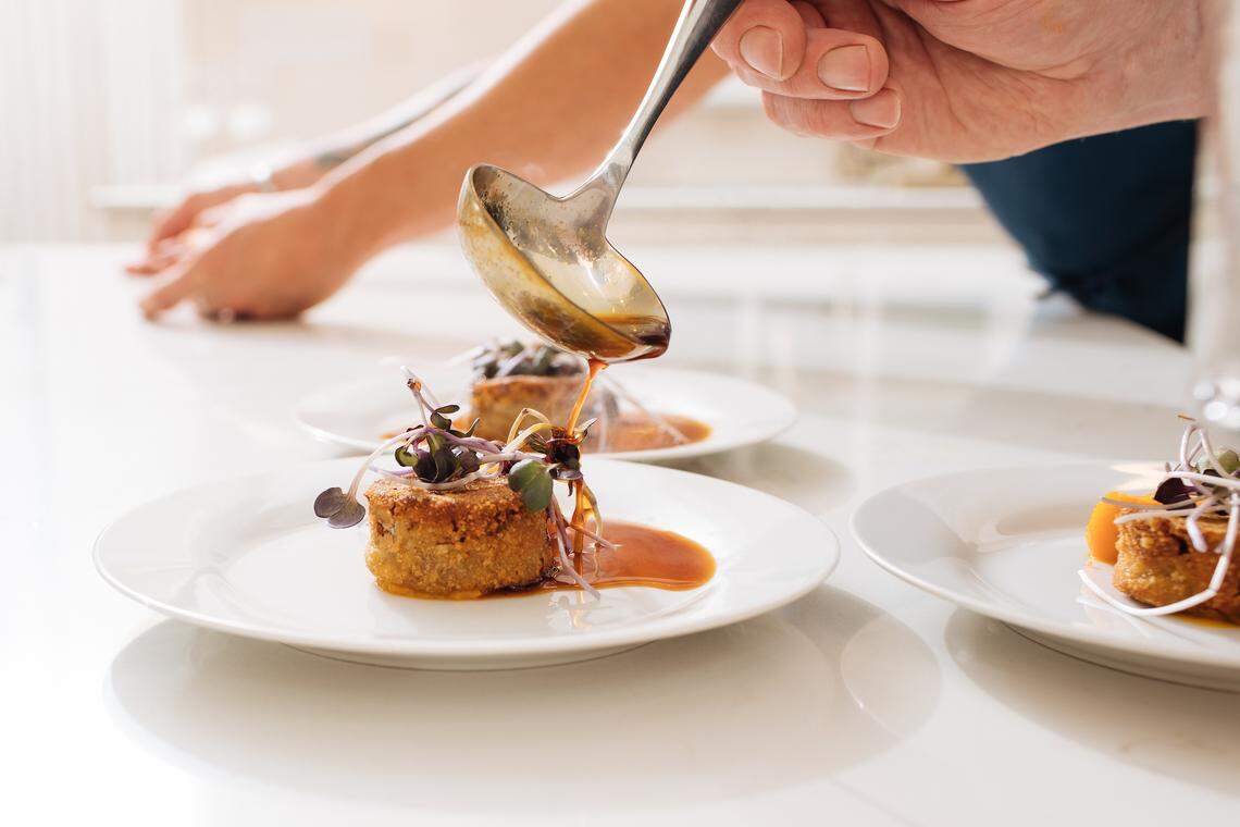 A close-up, brightly lit shot of a chef’s hand using a metal ladle to drizzle a glossy, amber-colored sauce over a small, circular, breaded savory cake.