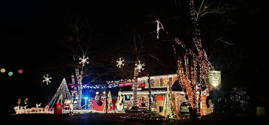 A highly dense and complex holiday light display covering the front of a home at night. The scene includes massive light-wrapped trees, an illuminated sleigh and reindeer, large light-up snowflakes suspended from the trees, and a sign asking for donations to St. Jude’s Children’s Hospital.