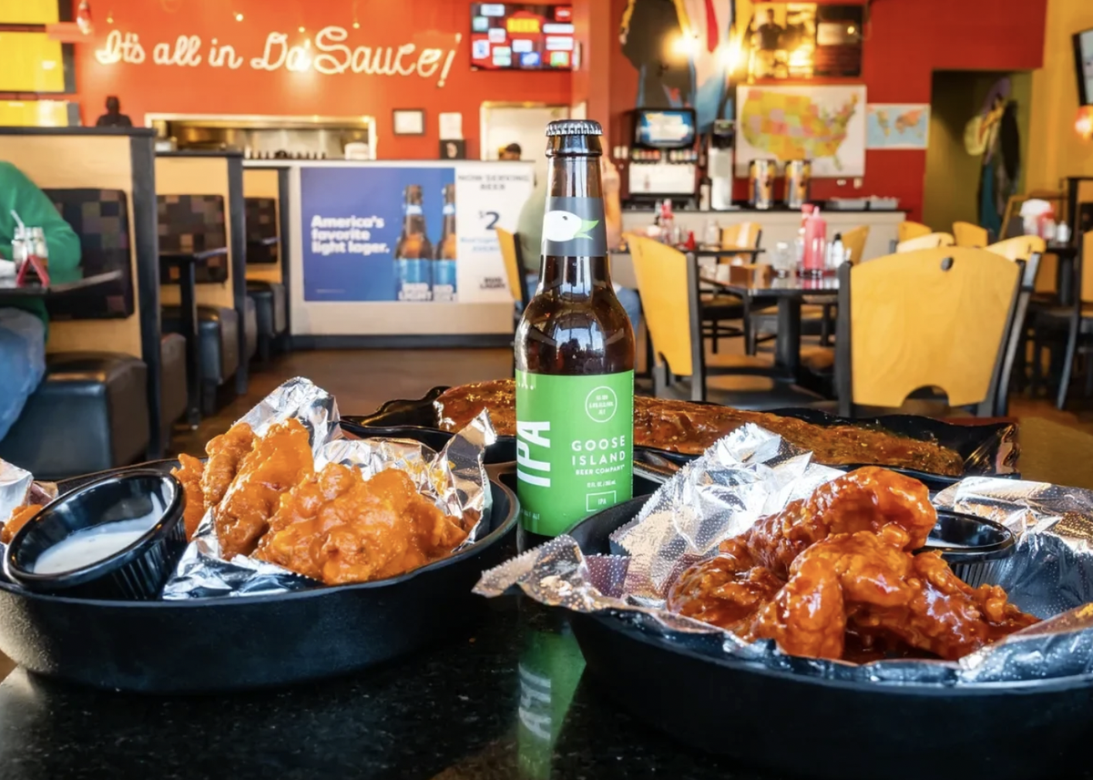 A close-up, low-angle shot of a dark wooden table topped with several baskets of chicken wings coated in bright orange sauce. A green glass beer bottle stands in the center of the frame, with the blurred background showing the red walls and yellow seating of the restaurant dining area.