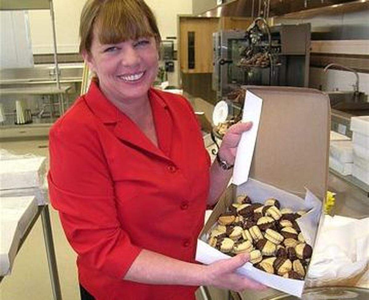 A woman smiles in a commercial kitchen. She is wearing a red collared shirt and holding a white cardboard box open to the camera, revealing it is full of numerous small, decorative sandwich cookies dipped in chocolate.