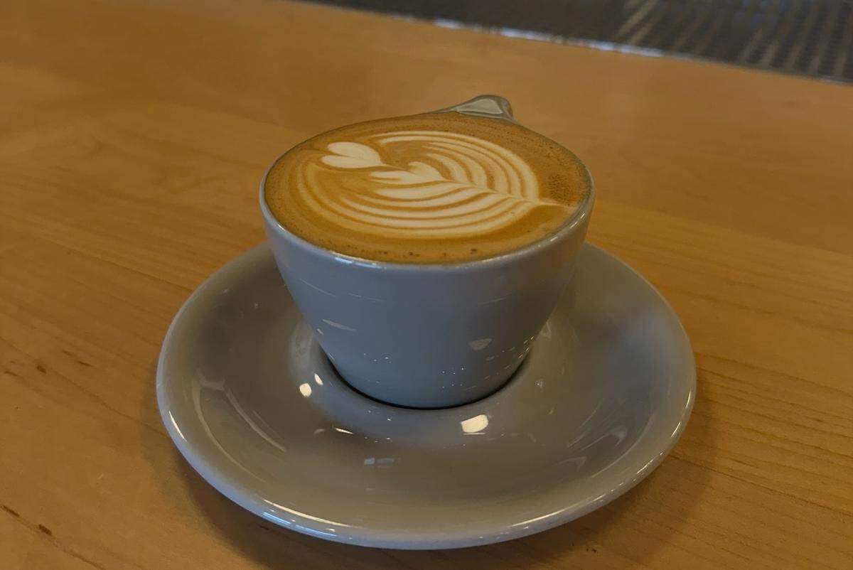 A close-up shot of a latte served in a light gray cup with a matching saucer, resting on a wooden bar. The coffee is topped with intricate, layered latte art in the shape of a rosetta. The coffee shop’s service area and a barista are blurred in the background.