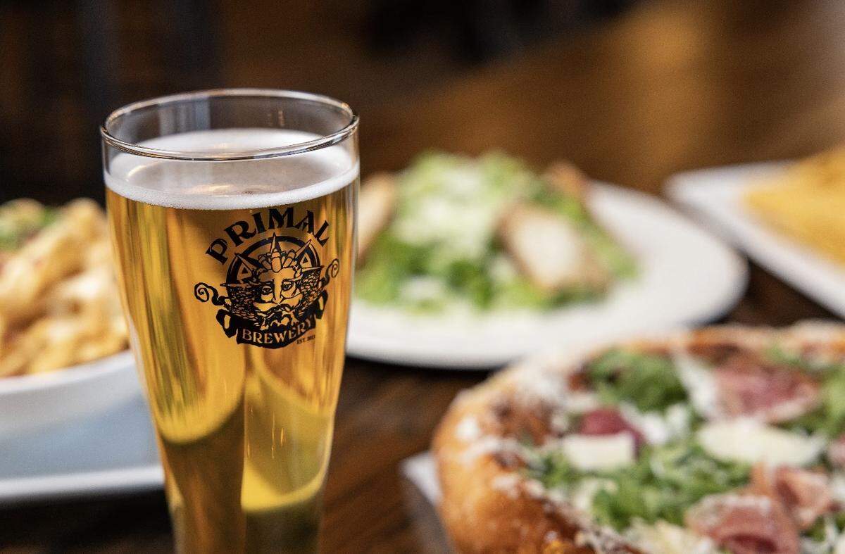 A close-up, shallow-depth-of-field shot of a tall glass of light lager featuring the Primal Brewery logo. In the blurred background, several food dishes are visible, including a fresh salad and a prosciutto-topped pizza.