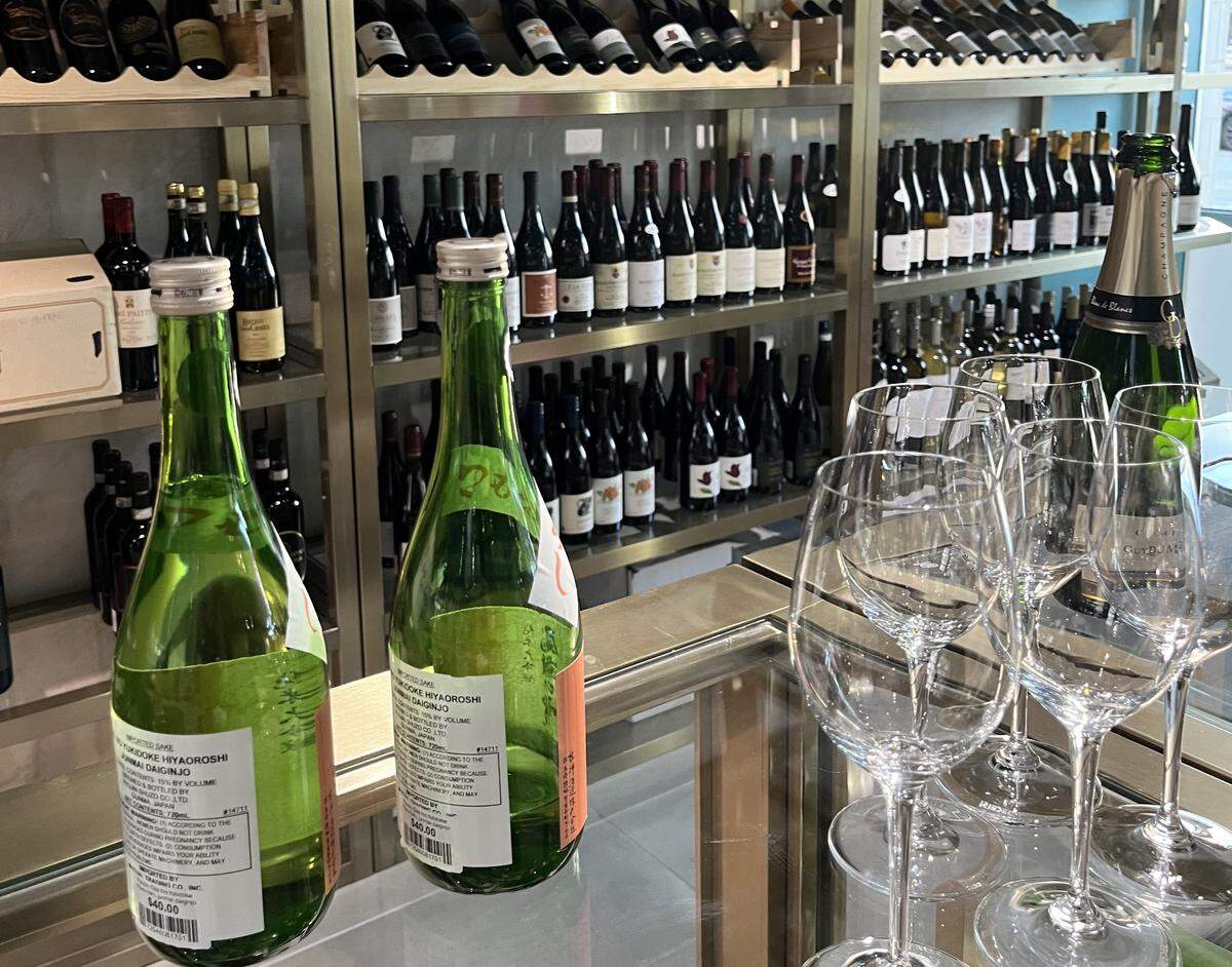 A close-up shot in a wine shop, focusing on a countertop with two green bottles of sake and a row of empty wine glasses. One of the sake bottles has a price tag that reads “$40.00”. In the background, metal shelves are densely packed with a wide variety of wine bottles.
