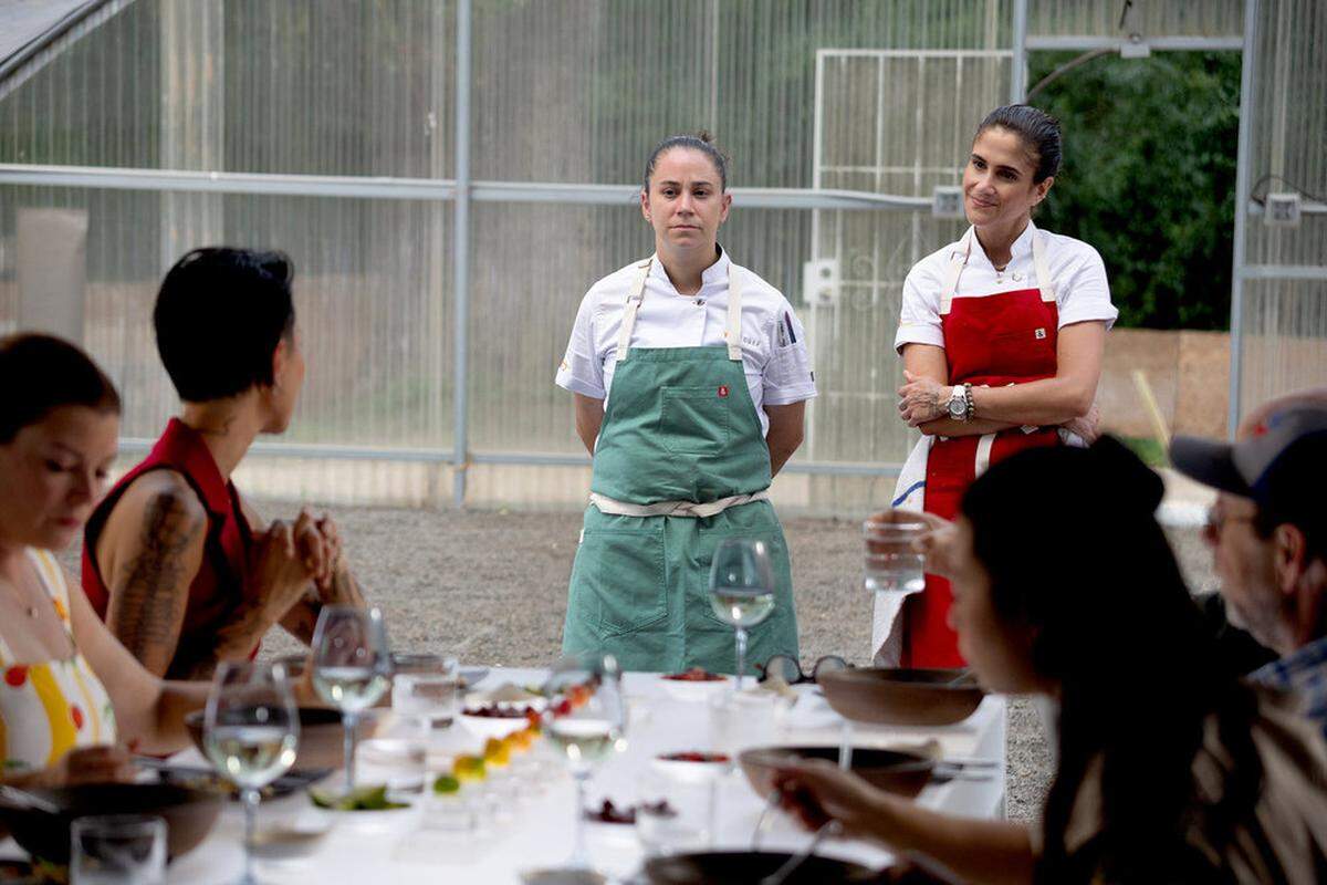 Two chefs, one in a green apron and one in a red apron, stand at the head of a long dining table inside a greenhouse-style structure. The chef in green stands with hands tucked behind, looking forward with a serious expression, while the chef in red leans slightly with arms crossed, looking toward the diners with a faint smile.
