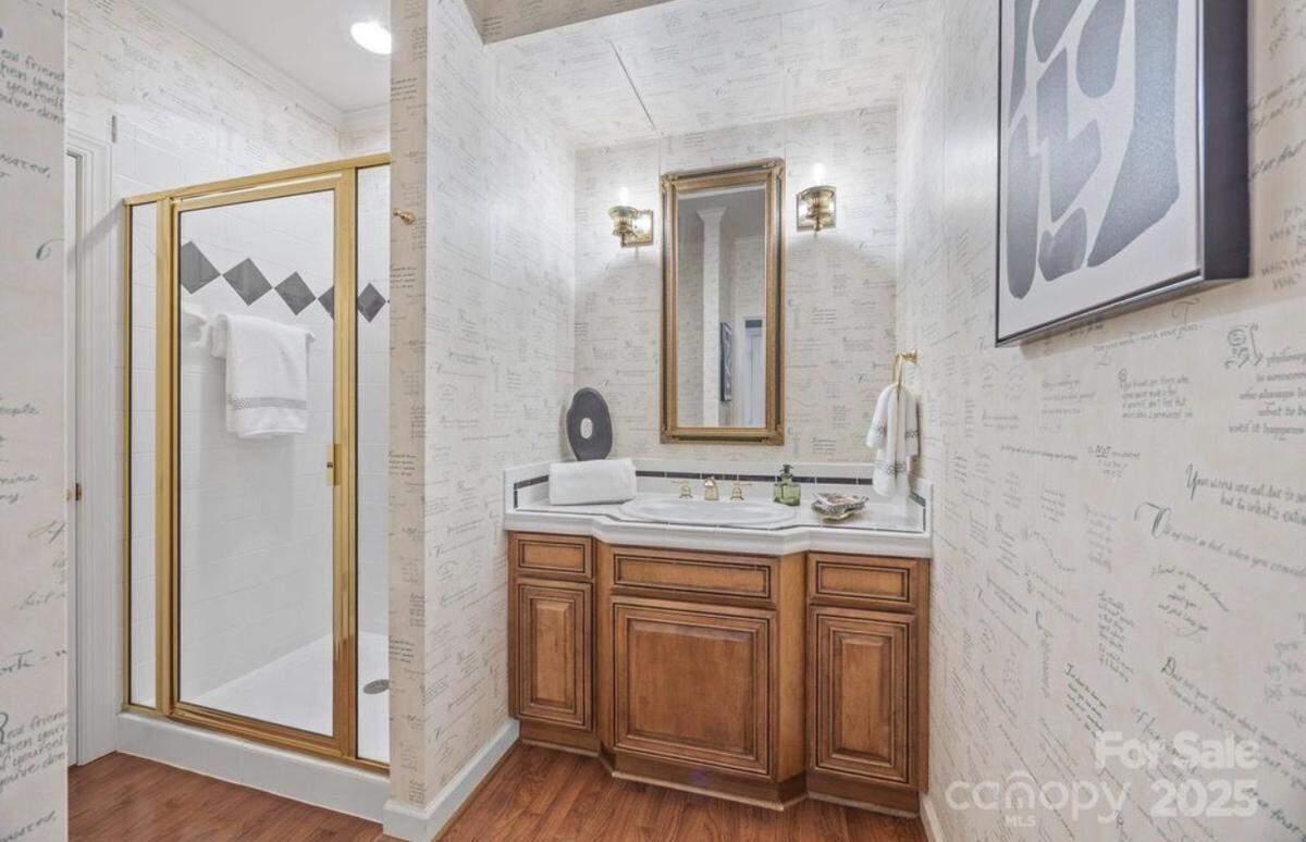 A bathroom interior featuring light walls covered in script wallpaper, a dark wood vanity with a white countertop and double-basin sink, and a framed mirror flanked by two sconce lights. To the left is a walk-in shower with a glass and gold-framed door, and to the right, a large framed black and white piece of art hangs over the wallpaper.