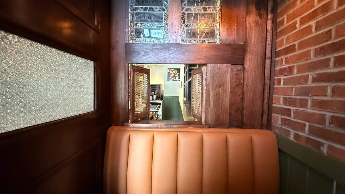 A close-up shot of a unique architectural detail inside the pub. Behind a tan leather booth, a wooden partition features etched glass panels with religious-style lettering and a small, open wooden “pass-through” window with decorative metal mesh. Through the window, a glimpse of the bar area and another piece of modern art can be seen. To the right, a textured red brick wall adds to the rustic atmosphere.