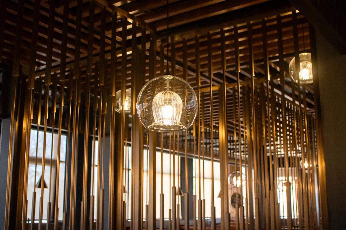 A close-up of modern restaurant ceiling decor, featuring a grid of wooden slats and several clear glass globe pendant lights. The lights cast a warm glow on the interior, and large windows are visible in the background.