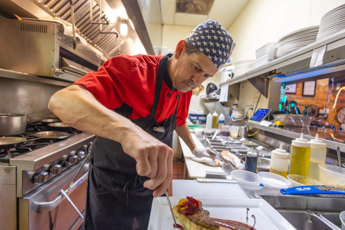A medium shot of a chef in a professional kitchen, wearing a red short-sleeved chef’s coat, a black apron, and a bandana with a blue and white star pattern, leaning over a stainless steel prep station, using a thin metal tool to carefully plate a gourmet dish on a rectangular white plate. The background shows the functional details of the kitchen, including a large industrial stove, ventilation hoods, and various prep containers and squeeze bottles.