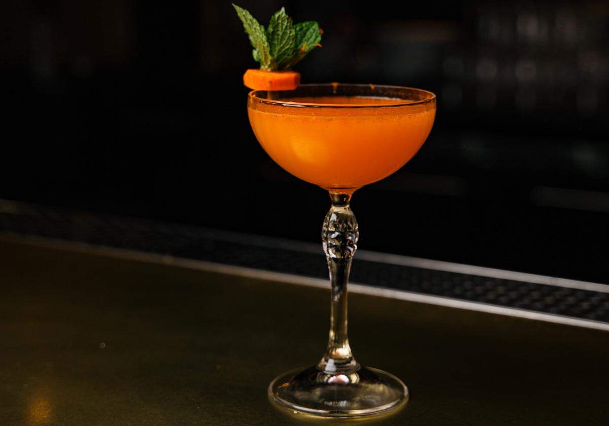 A striking, bright orange cocktail glows in a dimly lit bar. Served in a classic coupe glass with an ornate stem, the drink is simply garnished with a fresh sprig of mint and a round carrot slice, hinting at its ingredients. The lighting is focused and dramatic, creating a sophisticated mood.