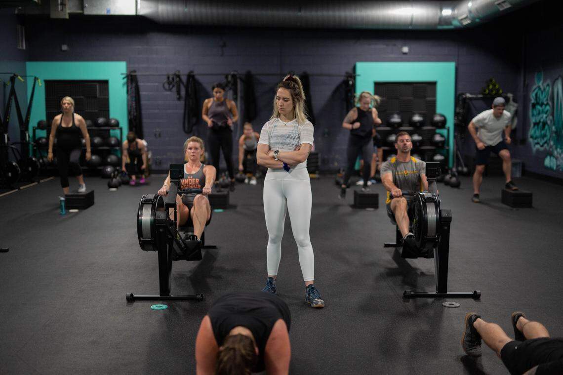 A fitness instructor in white athletic wear stands with arms crossed in the center of a gym. Several people are engaged in a workout, with two individuals using rowing machines and others using step boxes in a large, open space with dark walls and teal accents.