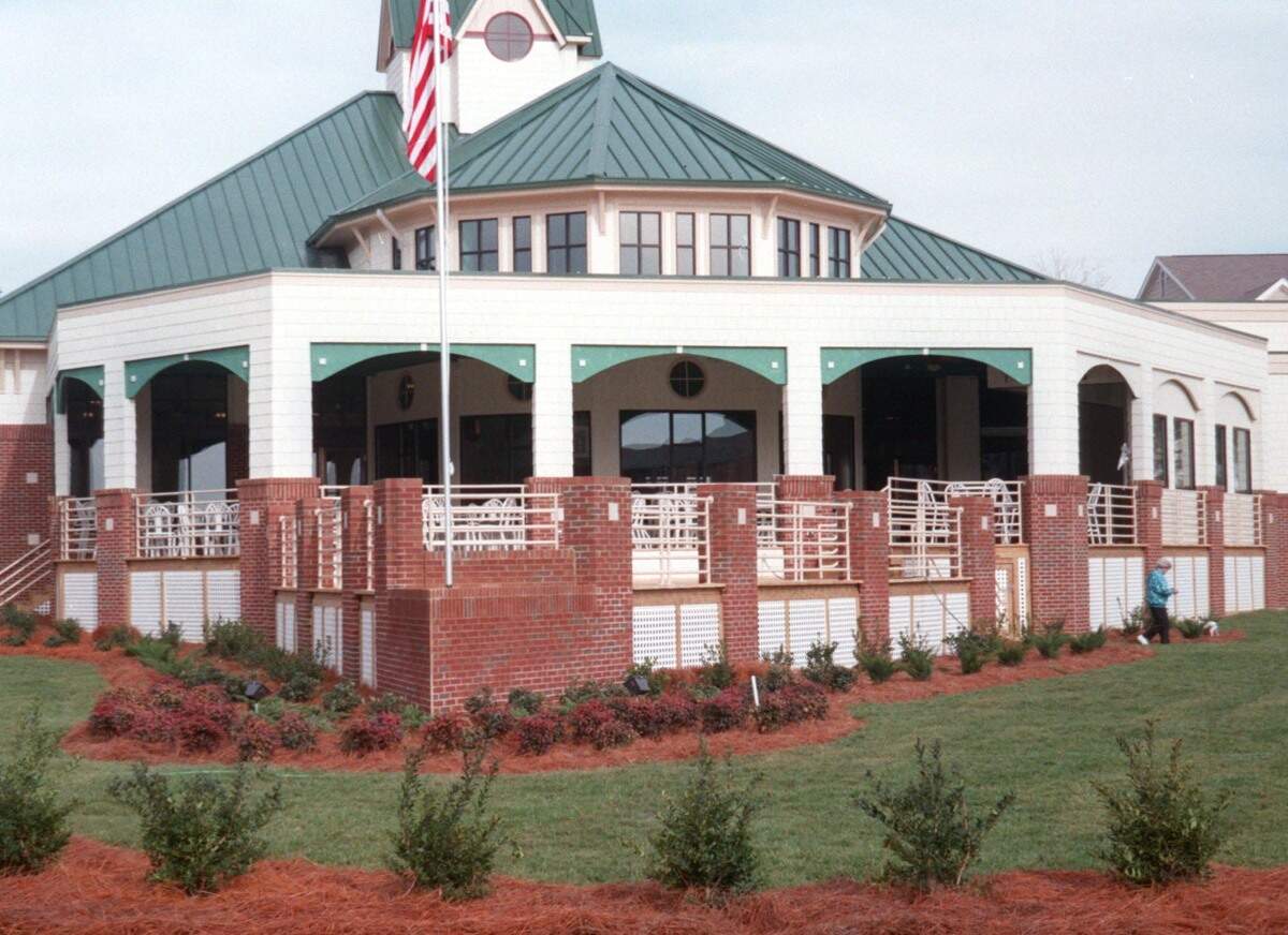 A wide, daytime shot of a large, octagonal building with a prominent green metal roof, a cupola at the peak, and white exterior walls. The building features an elevated outdoor patio surrounded by a red brick and white-railed perimeter wall. Red mulch flower beds with low green shrubs line the base of the structure, an American flag flies on a tall flagpole out front, and a person walks a small dog on the green lawn to the right.